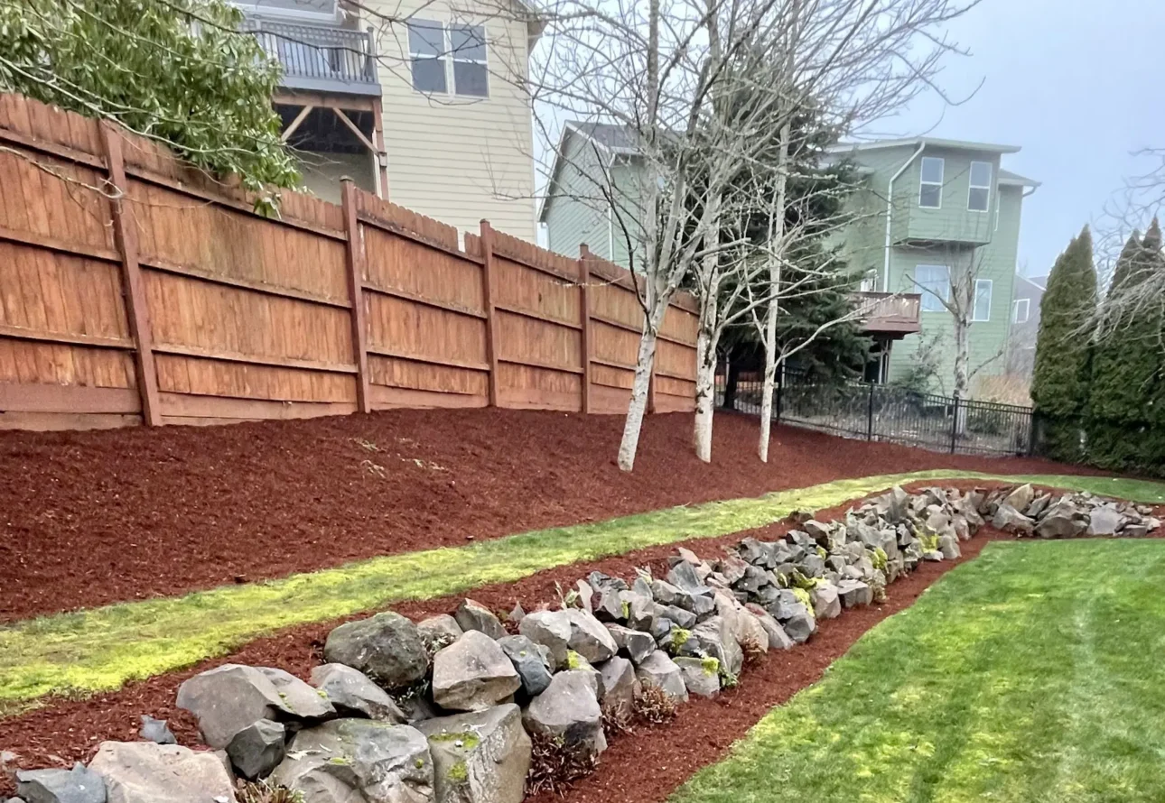 Sloped backyard landscaping with mulch beds, stone drainage channel, and wood privacy fence