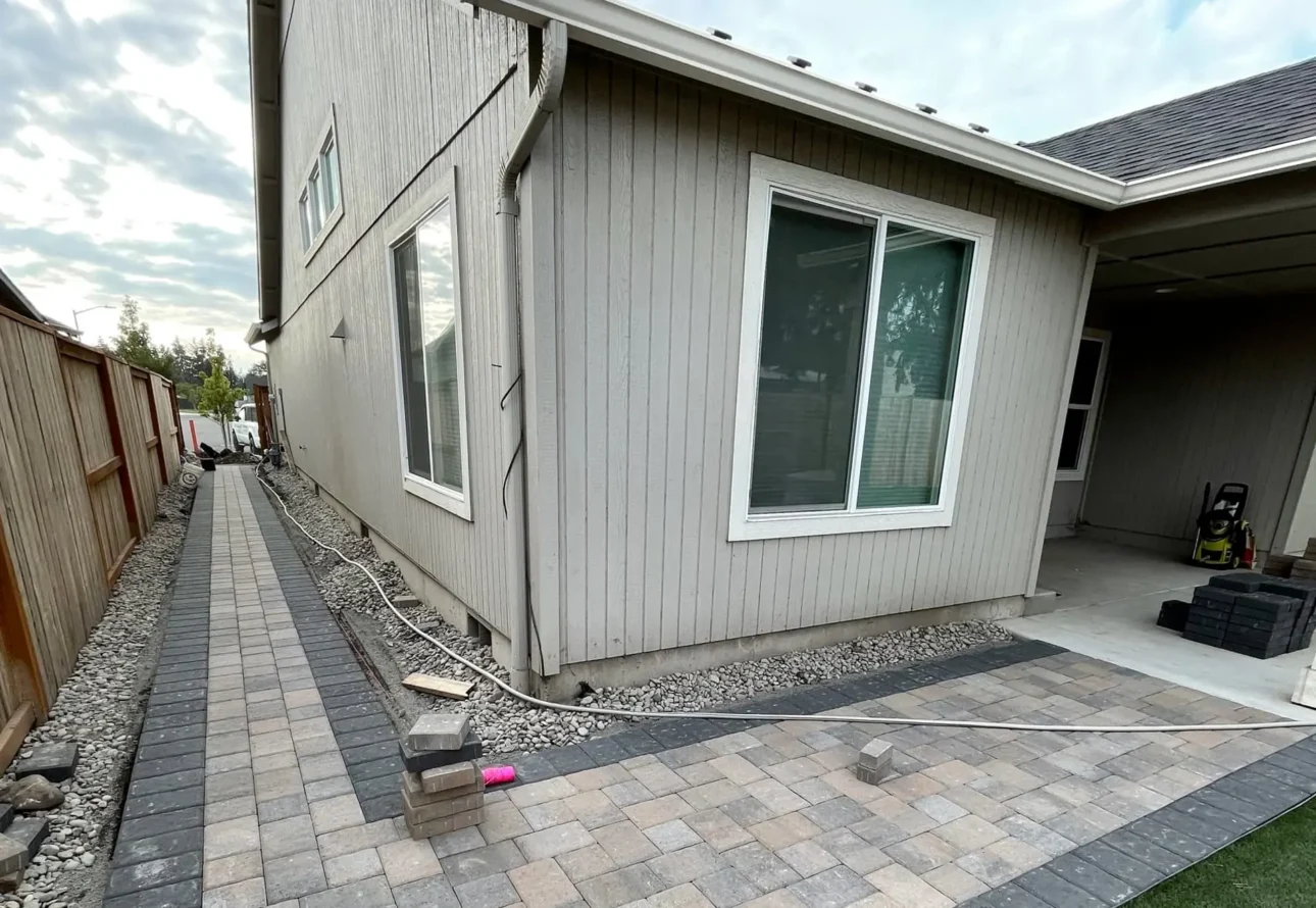 New paver patio and walkway installation in progress along a residential home with stone edging and gravel base